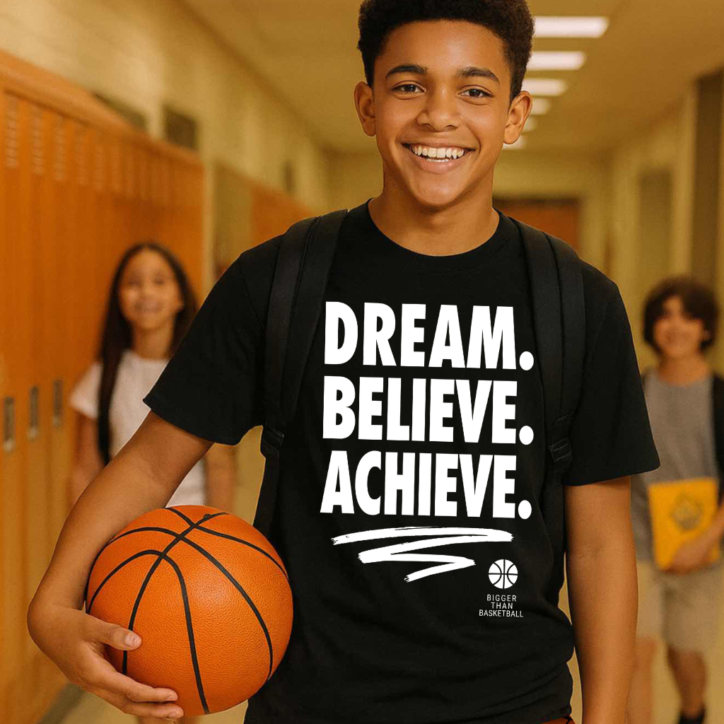 Young boy in a hallway holding a basketball, wearing a black t-shirt with motivational text and orange shorts.