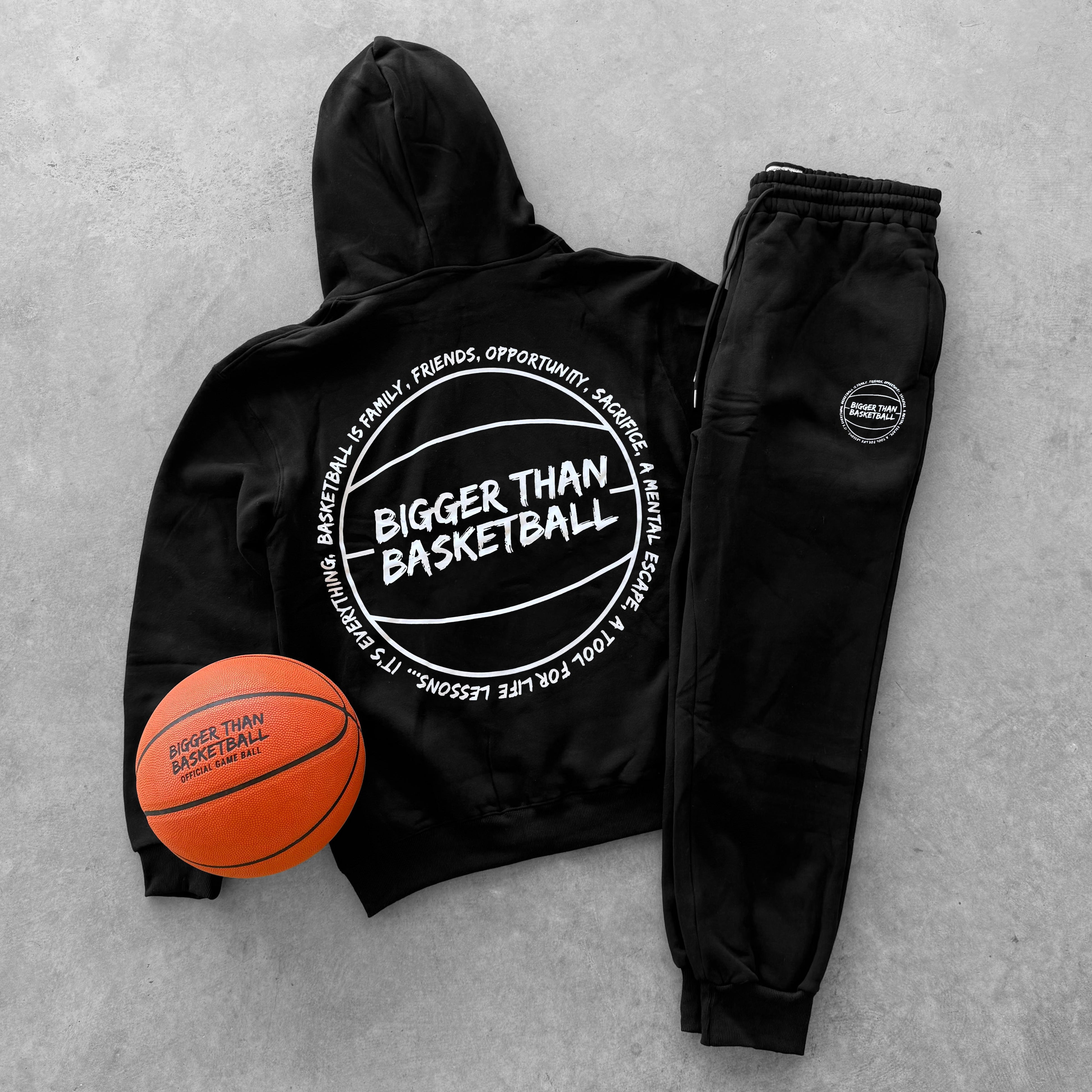 Black hoodie and jogger set with 'Bigger Than Basketball' text, accompanied by a basketball on a gray concrete background.